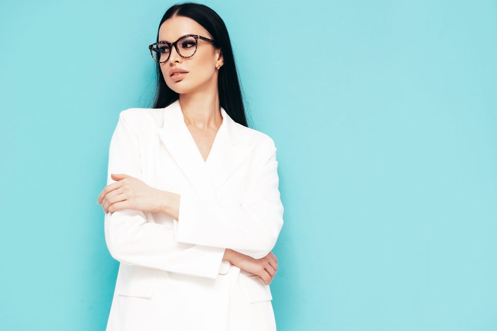 High fashion portrait of young beautiful brunette woman wearing nice trendy white suit. Sexy fashion model posing in studio. Fashionable female isolated on blue. Full length. In spectacles